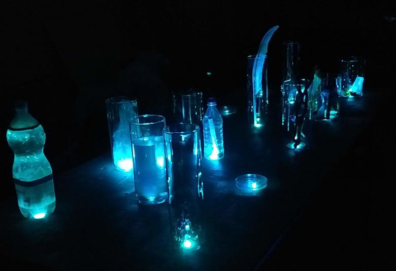 Kuvassa on tummalla pohjalla sinertäviä muovipulloja, vesilaseja ja muita esineitä.