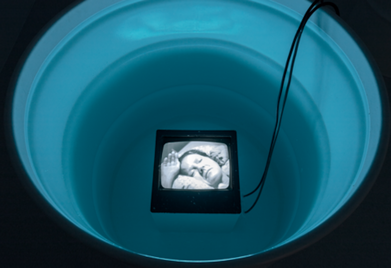 Kuvassa on musta vatimainen säiliö, jonka pohjalla on lepäävän naisen kuva.