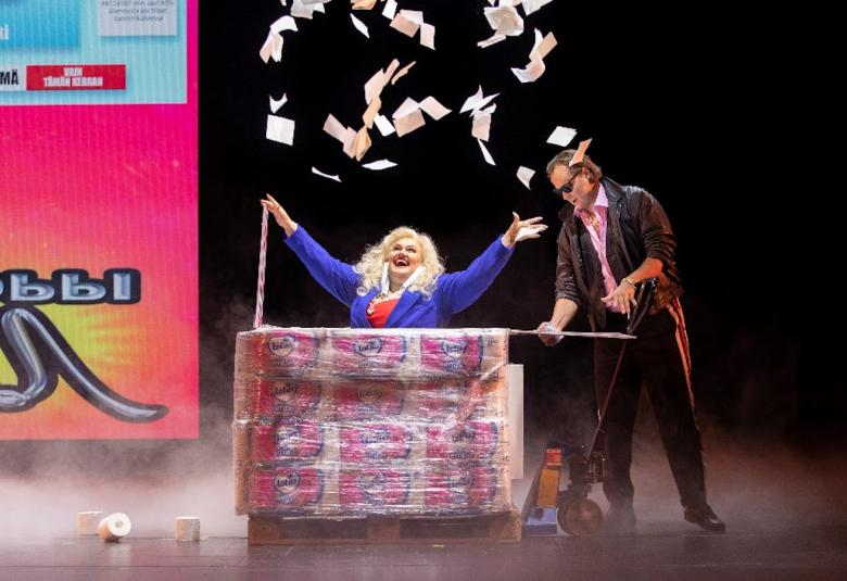 Kuvassa on vaaleahiuksinen nainen pöydän ääressä heitellen papereita ja vierellä on tummapukuinen mies. Seinällä on oikealla värikäs iso juliste.