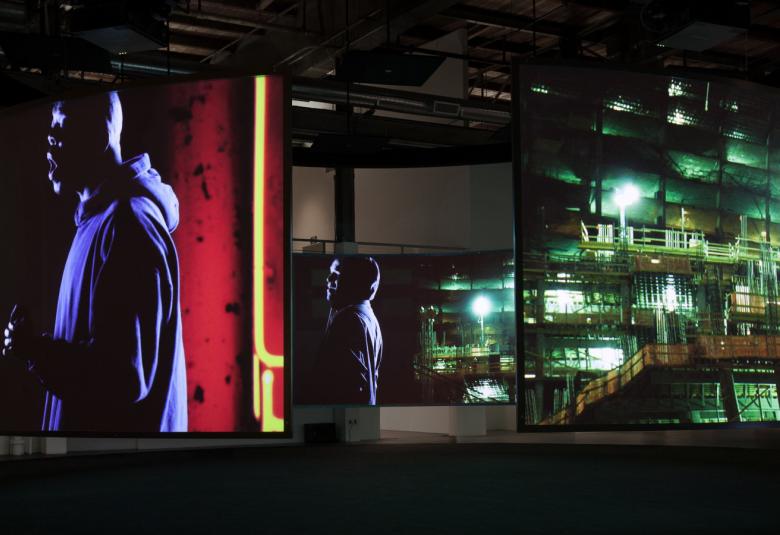 Kuvassa on Doug Aitkenin teos, jossa etualalla seisoo mies hupparissa ja vieressä on pienempi kuva miehestä. Molemmat ovat puoliksi selin. Taustalla näkyy yövalaistua modernin lasi-ikkunaisen rakennuksen seinää.