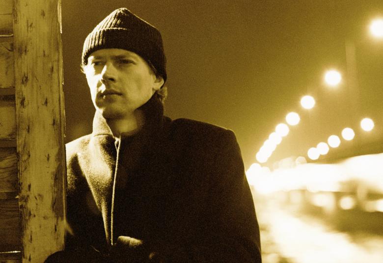 Kuvassa on Ilkka Remes musta-valkoisessa kuvassa pipo päässään vasemmalla katsomassa pois päin kuvasta ja taustallaan katunäkymää.