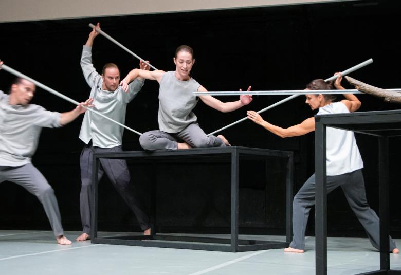 Kuvassa ovat tanssijat pitävät pitkiä keppejä kädessään ja ovat tanssimassa pöytien päällä ja vieressä vaaleissa asuissa.