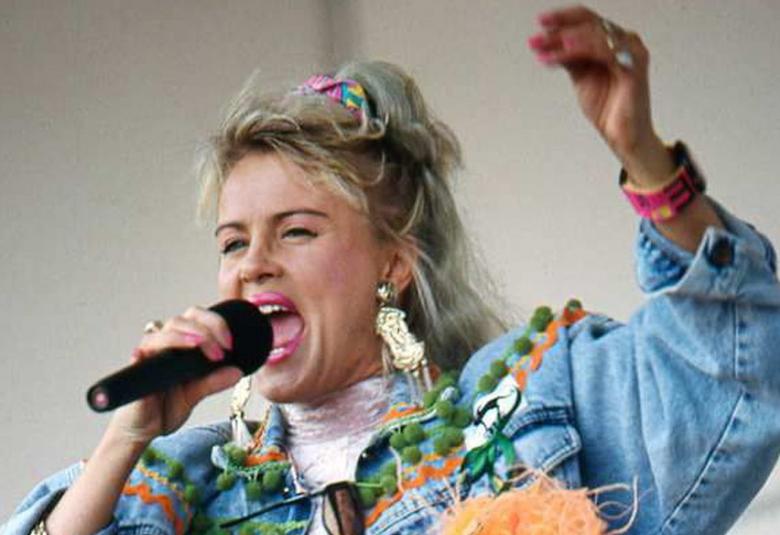 Kuvassa on Kikka esiintymässä mikki kädessään ja vaalean sinisessä farkkupusakassa. Kuva on puolivartalokuva.