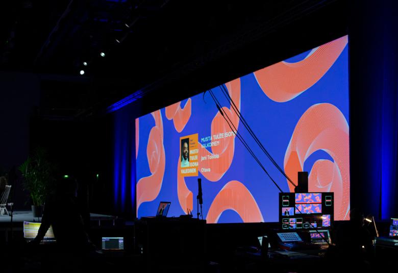 Kuvasssa on Helsingin kirjamessujen esiintymislava ja siinä iso skriini, josta näkyy lavalla oleva puhuja.