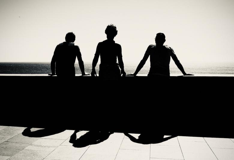 Kuvassa on kolme miestä kuvattuna takaa päin ja he katselevat merelle betonisen kaiteen äärellä. Kuva on musta-valkoinen.