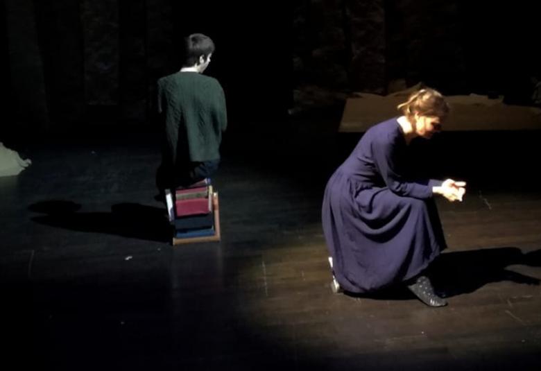 Kuvassa näyttelijät istuvat selät melkein vastakkain tummissa asuissa ja näyttämön valaistus on myös tumma.  