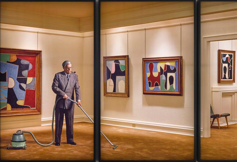 Kuvassa on mies puvussa imuroimassa taidegalleriassa.  Seinillä on modernia taidetta ja vasemmassa reunassa on nainen katselemassa tauluja.