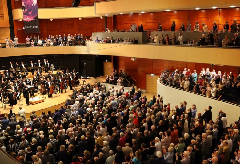 Kuva on Sibelius-talon konserttisalin konsertista, jossa on orkesteri kiittämässä ja sali täynnä yleisöä. 