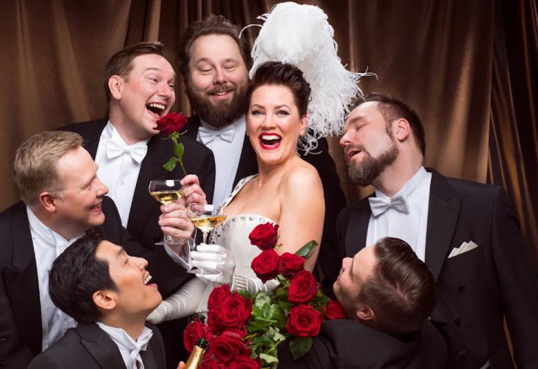 Kuvassa on keskellä Mari Palo ja ympärillä kuusi miesesiintyjää ruusujen kera.