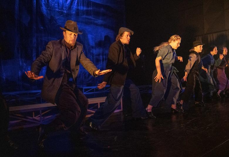 Kuvassa ovat tanssijat rivissä näyttämöllä vauhdikkaassa liikkeessä.  Heillä on harmahtavat puvut päällään.