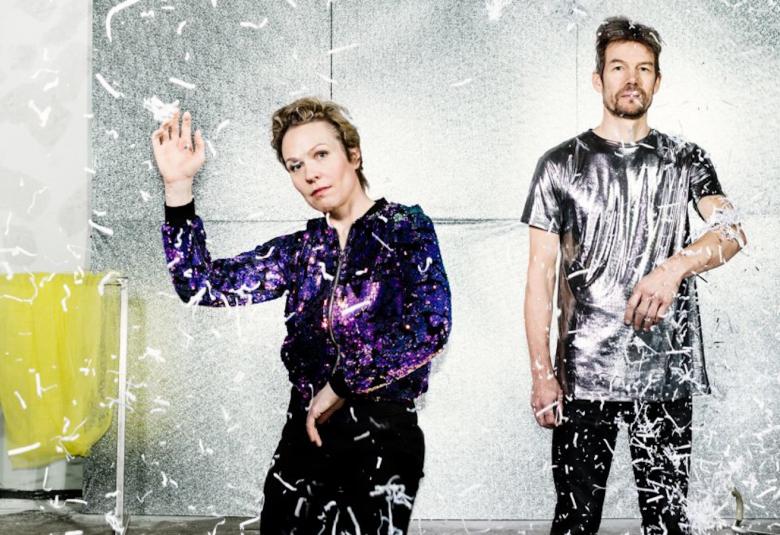 Kuvassa ovat seisomassa Maija Hirvanen ja Juha Valkeapää tilassa, jonka taustana on harmaa taulu ja vasemmalla roikkumassa vihertävä kangas.  Ilmassa sataa paperisuikaleita.