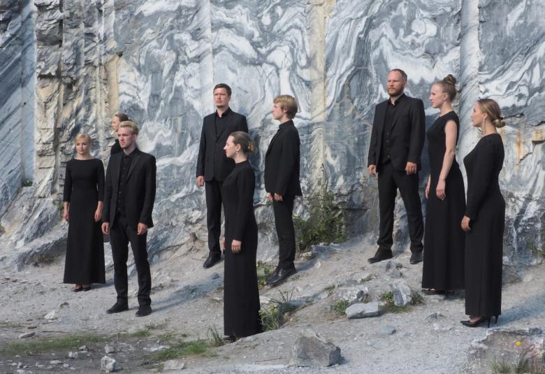 Kuvassa on näyttelijöitä mustissa puvuissa kulisseissa ja takana harmahtava metsä.