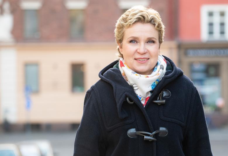 Kuvassa on Sirpa Kähönen duffelitakki päällä ja hänellä on vaalea huivi kaulalla. Taustalla on kellertävä vanha rakennus ja vasemmalla näkyy Suomen lippu.