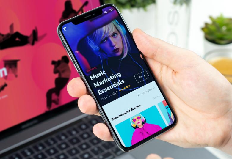 Kuvassa on näyttö pöydällä ja henkilön kädessä kännykkä, josta näkyy näyttö.