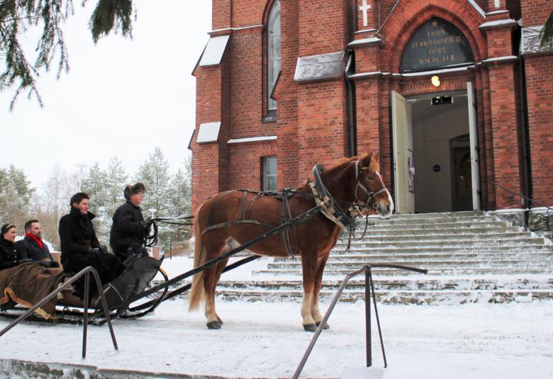 Kuvassa on hevosen vetämässä reessä Jussi Merikanto, Jussi Makkonen ja Nazig Azezian Nurmeksen punatiilisen kirkon edessä.