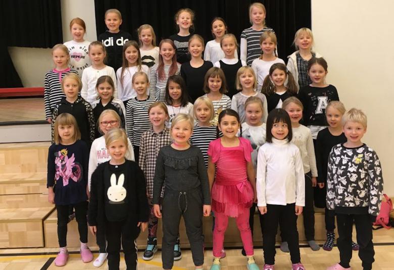 Kuvassa on Jyväskylän normaalikoulun pienten kuoro harjoituksissa ja he ovat neljässä rivissä omissa vaatteissaan.