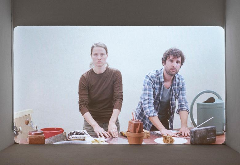 Kuvassa on nainen ja mies seisomassa pöydän ääressä edessään saviruukkuja ja tavaroita, mm. matkaradio. Kuva on tehty kuvaruutumaiseksi.