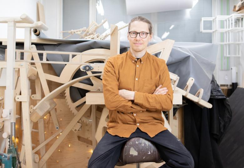 Kuvassa on Petri Eskelisen puolivartalokuva ja hän on keskellä työhuonettaan puisten materiaaliensa keskellä.