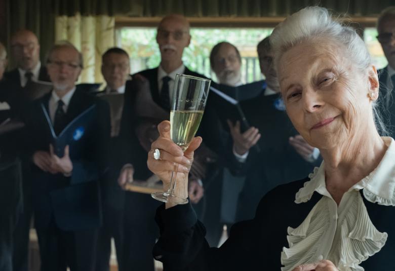 Kuvassa on Seela Sella kuohuviinilasi kädessä. Kuva on puolivartalokuva ja taustalla näkyy himmeästi miesryhmä.