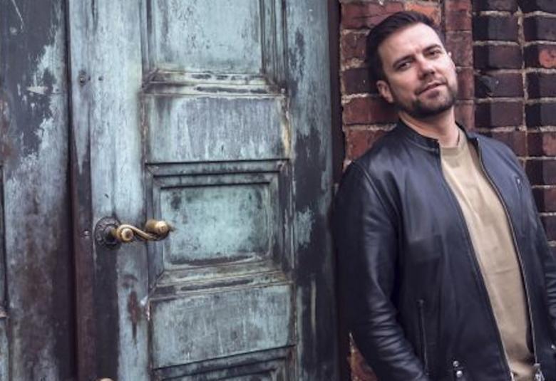 Kuvassa Antti Ketonen nojaa tummahkoon tiiliseinään ja vieressä on harmaa kulunut kaksoisovi.