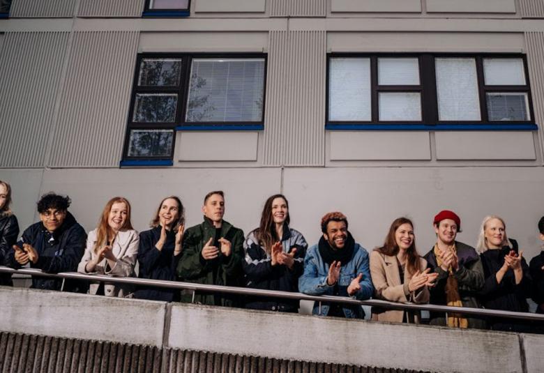Kuvassa on teatterikorkeakoulun opiskelijoita rivissä kaiteen takana rampissa ja takana on harmaapintainen kerrostalo ikkunoineen.