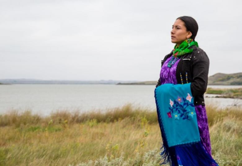 Kuvassa on Floris Ptesáŋ Huŋká katsomassa tasangolla avaraa maisemaa ja seisoo sivuttain traditionaalisessa tumman violetissa asussa.