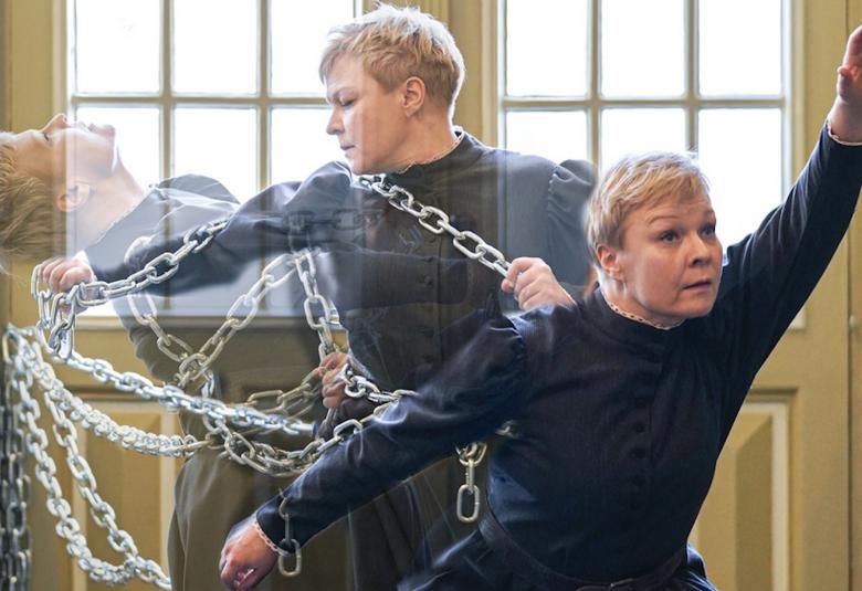 Kuvassa on tanssija, jonka ylävartalo heijastuu useana kuvana ja joka murtautuu kettingistä.  Taustalla on ruudukkoikkunaa.