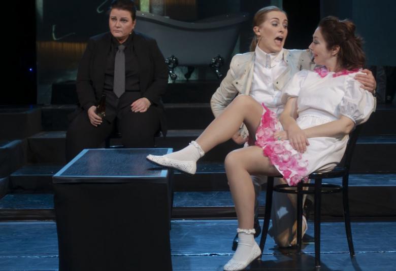 Kuvassa ovat etualalla vaaleissa vaatteissa kaksi naista, joista toinen laulaa ja taustalla on tummassa takissa ja kravaatissa mieheksi pukeutunut nainen.