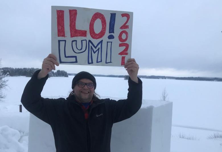 Kuvassa on Reko Mäki pitelemässä kädet ylhäällä kylttiä, jossa lukee Ilo!lumi 2021 ja taustalla on luminen järven selkä.