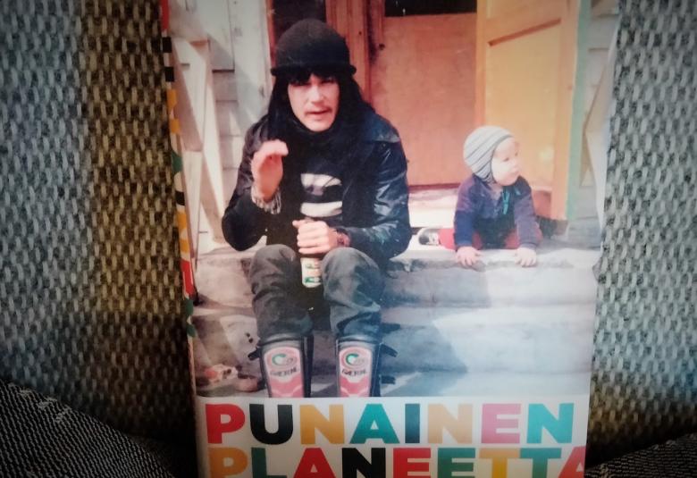 Kuvassa on talon portailla Jooonatan Tola ulkovaatteissa ja takana ryömii piene lapsi.  Alla likee Punainen planeetta isoin kirjaimin.