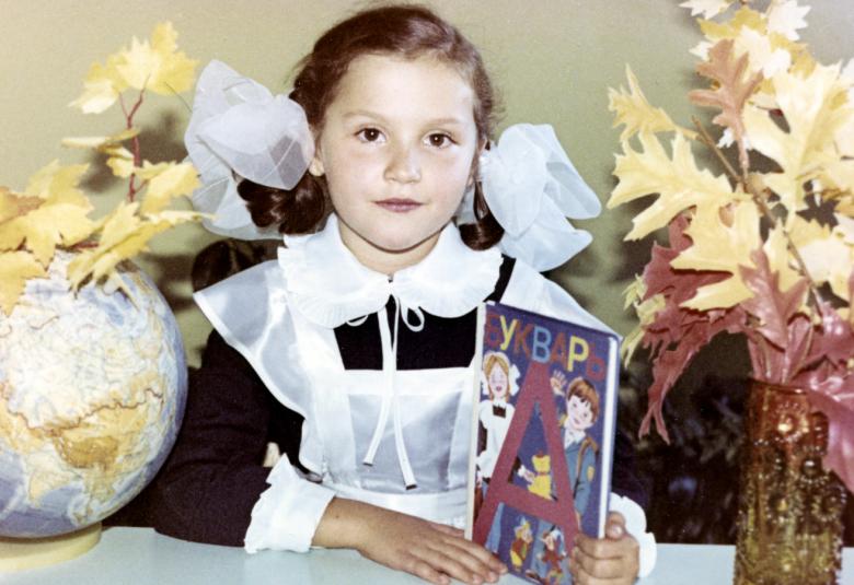 Kuvassa on Neuvostoliitossa asuva tyttö koulupuvussa isot rusetit päässa ja koulukirja kädessä.  Hän istuu valkoisen pöydän ääressä. 