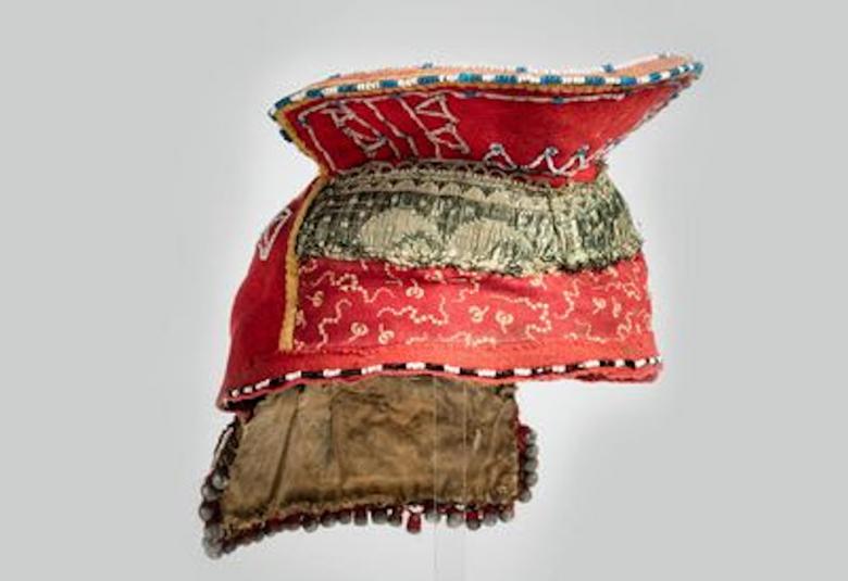 Kuvassa on kolttasaamelainen päähine, jossa on yläosassa ja keskellä ruskea punaista ja väleissä ja takana vihertävää kangasta.  Kuvioita on koko päähineessä.