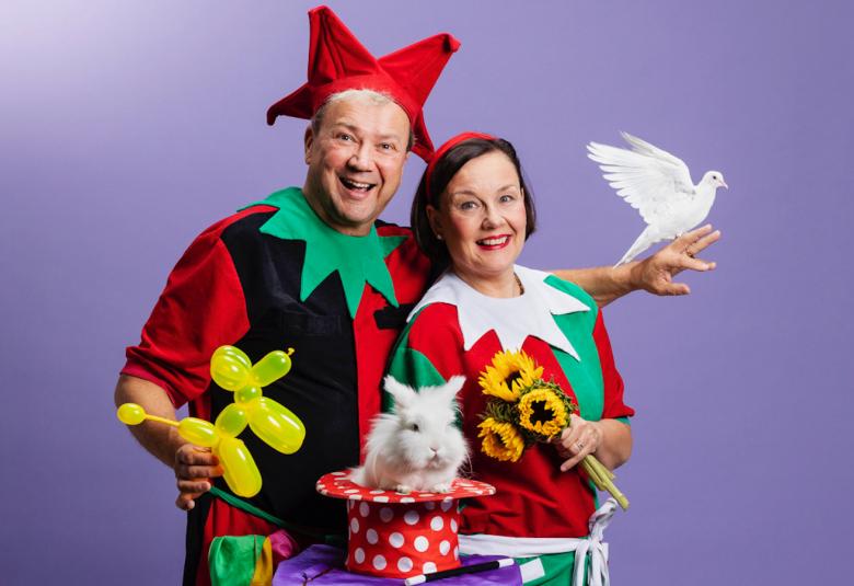 Kuvassa ovat Simo ja Kirsti Aalto värikkäissä asuissa ja Simo Aallolla on punainen neljäntuulen lakki päässään.  Edessä on pöytä, jossa on taikurin hattu, jossa istuu leikkikani.