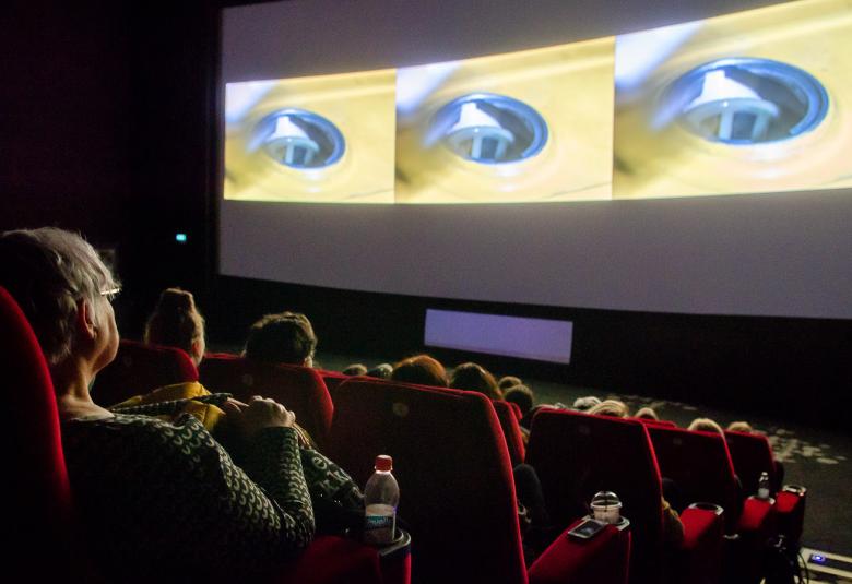 Kuvassa on elokuvateatteri, joka laskeutuu alaviistoon ja siinä on katsojia.  Valkokankaalla näkyy lieriöitä.