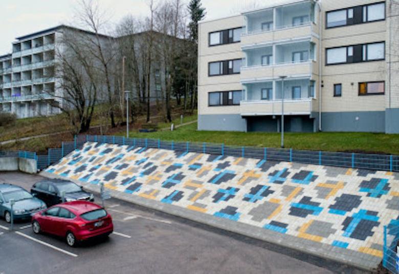 Kuvassa on parkkipaikka, jonka viereisellä penkereellä on mosaikkimainen laatta.  Värit ovat sinistä, vaalean keltaista, vaalean harmaata ja valkoista.