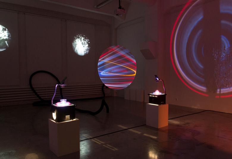 Kuvassa on pimeähkössä tilassa keskellä kaksi piirtoheitintä, joiden kannella on valossa pieni vesimalja.  Keskellä on pyöreä monivärivalo ja seinällä kaksi pyöreää mustavalkokuvaa.