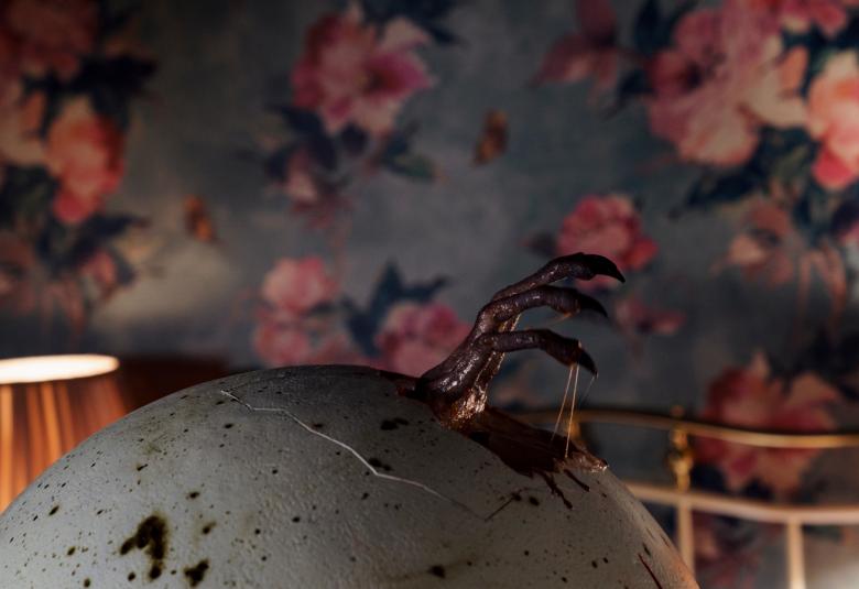 Kuvassa on suuri tumman vihreä muna ja siitä näkyy linnun musta jalka. Taustalla näkyy himmeästi punaruusukuvioista tapettia.