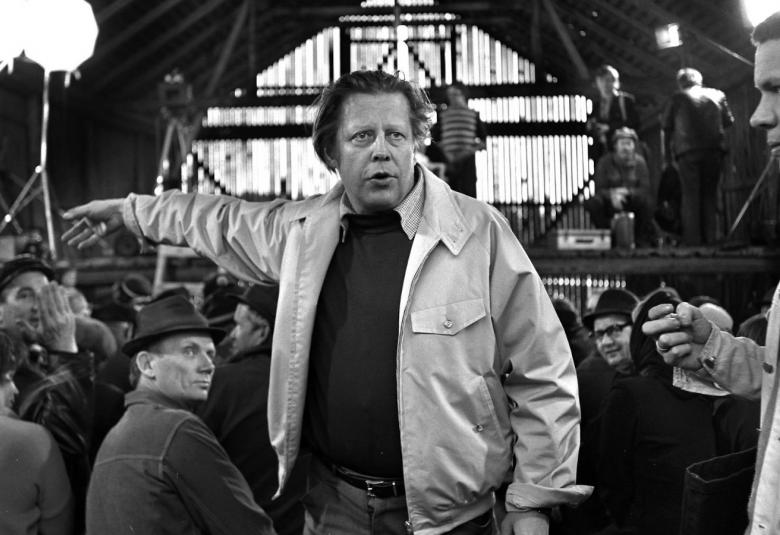 Kuvassa on keskellä Rauni Mollberg ohjaamassa Siunattu hulluus -elokuvaa.  Taustalla on miehiä istumassa penkeissä puisessa ladossa.