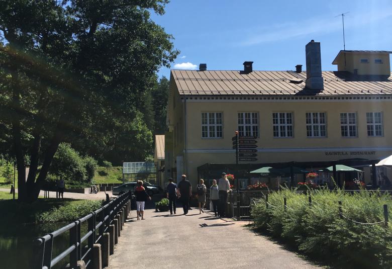 Kuvassa on kesäkuva Fiskarsista ja suuri keltainen vanha talo.  Ihmisiä on kulkemassa on kohti taloa hiekkakäytävää pitkin.