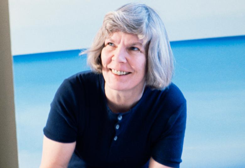 Kuvassa on Maija Isola lattialla polvillaan tekemässä luonnosta kankaasta ja hän katsoo hymyillen eteenpäin.