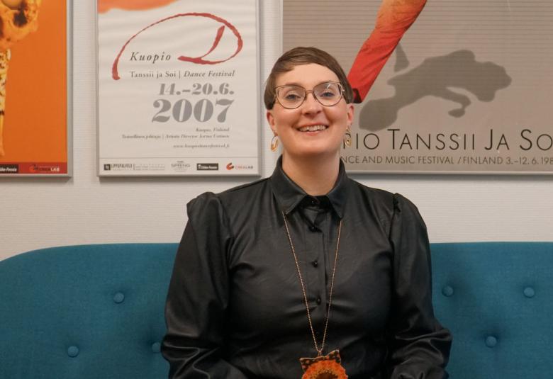 Kuvassa on Johanna Rajamäki istumassa sohvalla.  Taustalla näkyy Kuopio Tanssii ja Soi juliste ja muita julisteita.