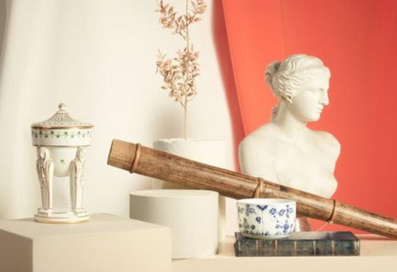 Kuvassa on vaalean pöydän ja jalustan päällä posliiniesineitä ja kaukoputki sekä klassinen naispatsas kasvoista.  Takana on vaalea kultaisella kukkakuviolla varustettu liina ja oikealla on ruosteen punaista seinää.