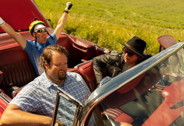 Kuvassa ovat punaisessa avoautossa etupenkillä ohjaajana Jaajo Linnoinmaa ja Sami Hedberg sekä takapenkillä Aku Hirviniemi kädet ojennettuina ylöspäin.  Kuva on otettu kesällä.