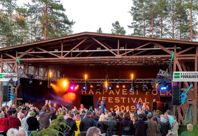 Kuvassa on puinen esiintymislava, jolla on esiintyvä bändi ja etualalla on yleisöä kesäisessä illassa.