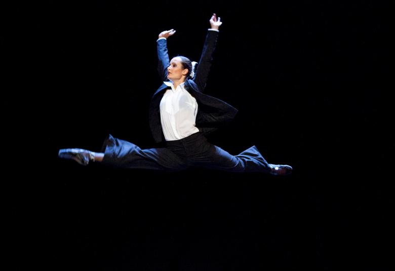 Kuvassa on balettitanssija Elza Leimane ilmassa spagaatissa ja hänellä on sininen puku ja valkoinen paitapusero päällään. Tausta on musta.