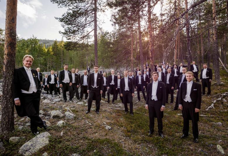 Kuvassa ovat Ylioppilaskunnan Laulajat metsässä esiintymisasuissaan frakeissa ja vasemmalla edessä on johtaja Pasi Hyökki seisomassa koivun vieressä. 