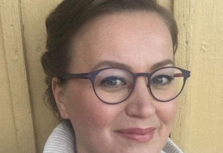 Kuvassa on Anna-Riikka Carlssonin kasvokuva.  Hänellä on tummat silmälasit ja vaalea pusero.