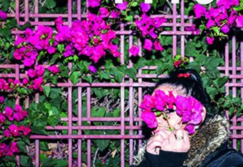 Kuvassa on punaista kukkaköynnöstä seinällä ja nainen oikealla seisomassa kasvot peitettynä punaiset kukat silmillään.