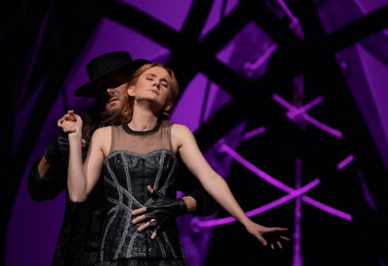 Kuvassa on tummalla näyttämöllä lierihattuinen mies, joka on vartalomyötäisessä siniharmaassa ja hihattomassa puvussa.