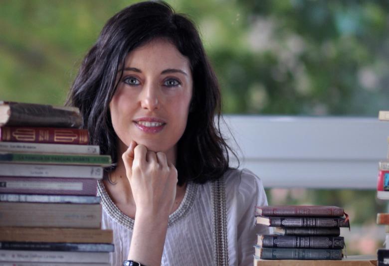 Kuvassa on Irene Vallejo istumassa kirjoituspöydän ääressä ja pöydällä on kirjapinoja.